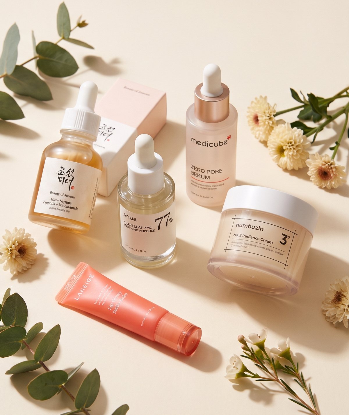 A selection of Korean beauty products, including a Beauty of Joseon glow serum, an Anua heartleaf ampoule, a medicube serum, a numbuzin cream and a Laneige lip balm, arranged with eucalyptus and dried flowers on a cream background.